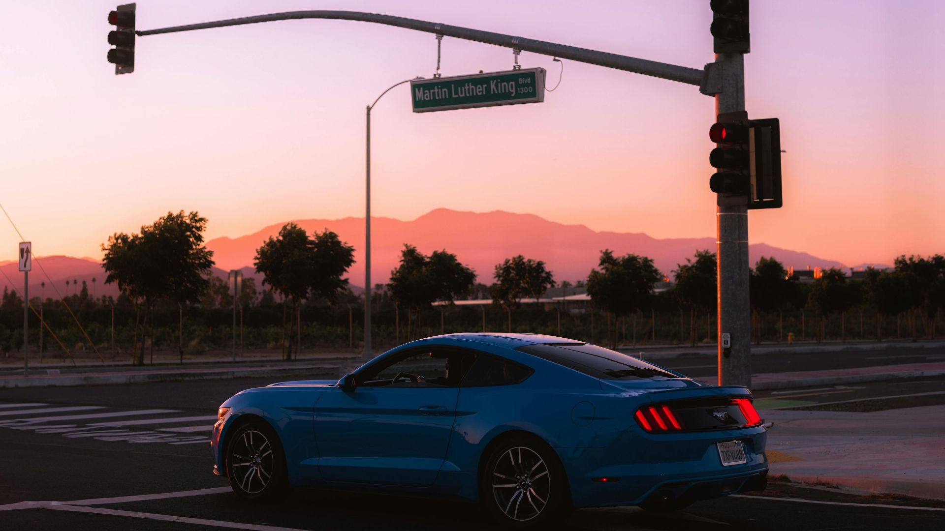 Blue sports car at a road intersection during sunset with a mountainous backdrop.