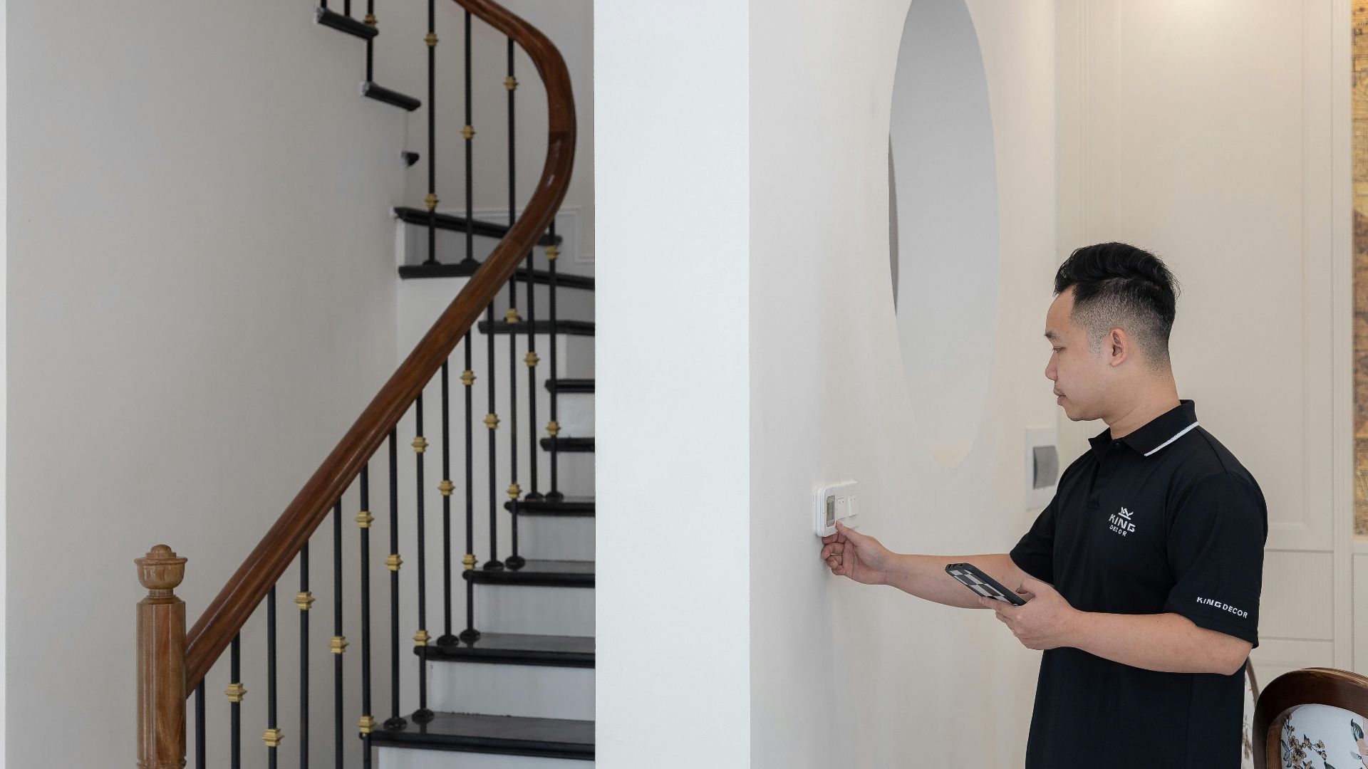 Man adjusting thermostat in stylish home with elegant staircase.