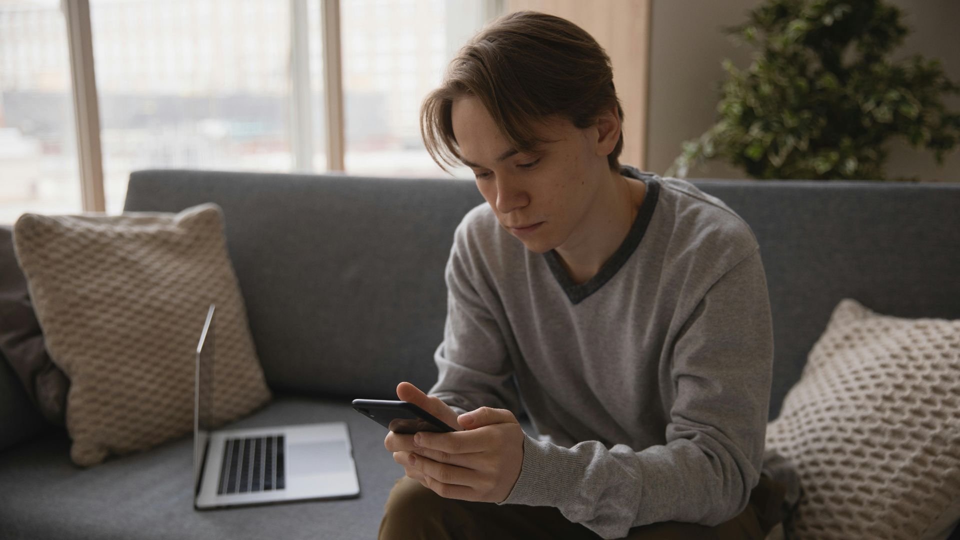 A young man sits on a couch using his smartphone with a laptop nearby. Modern technology indoors.