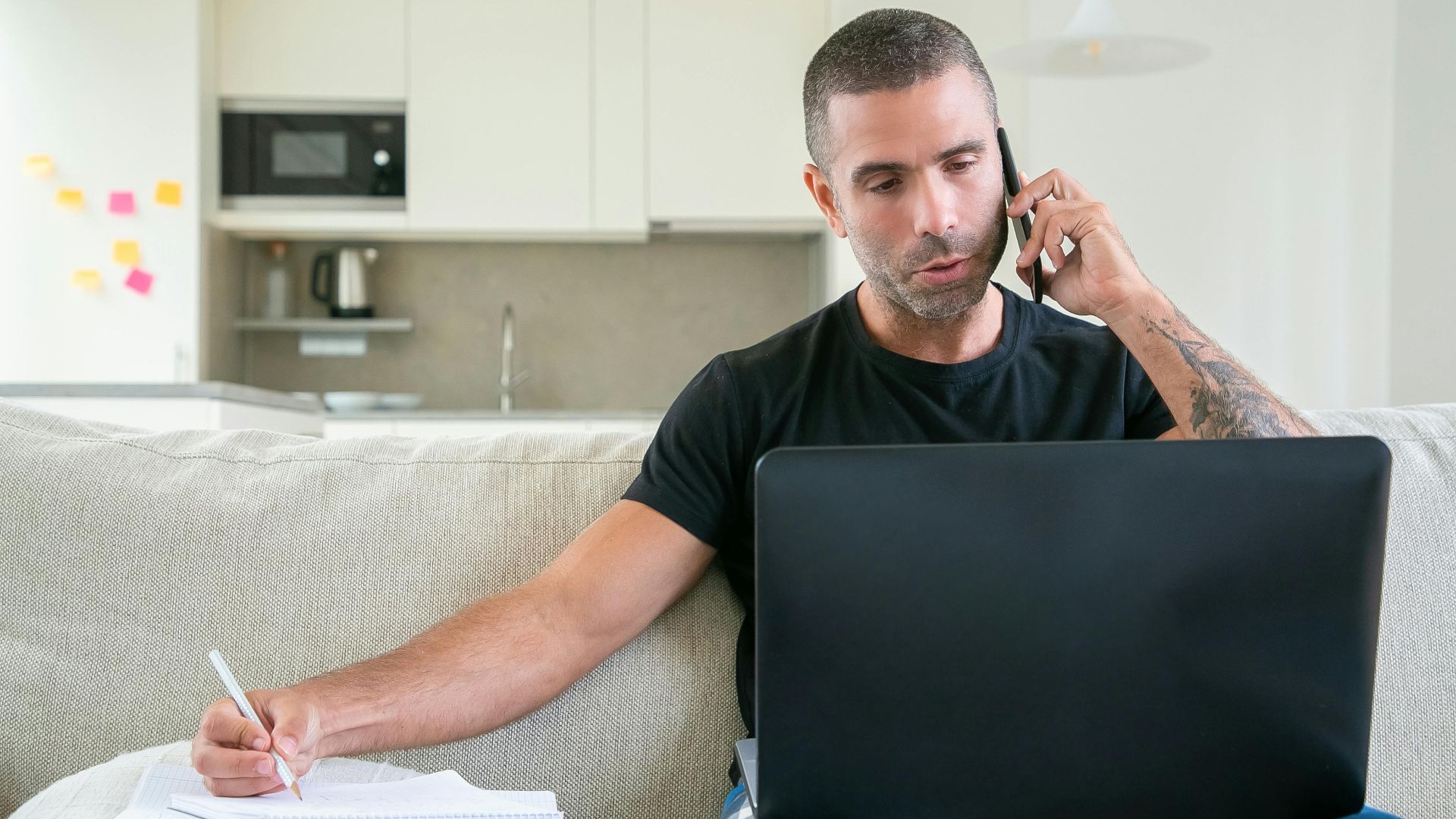 Man working from home using a laptop and smartphone while taking notes in a modern kitchen setting.