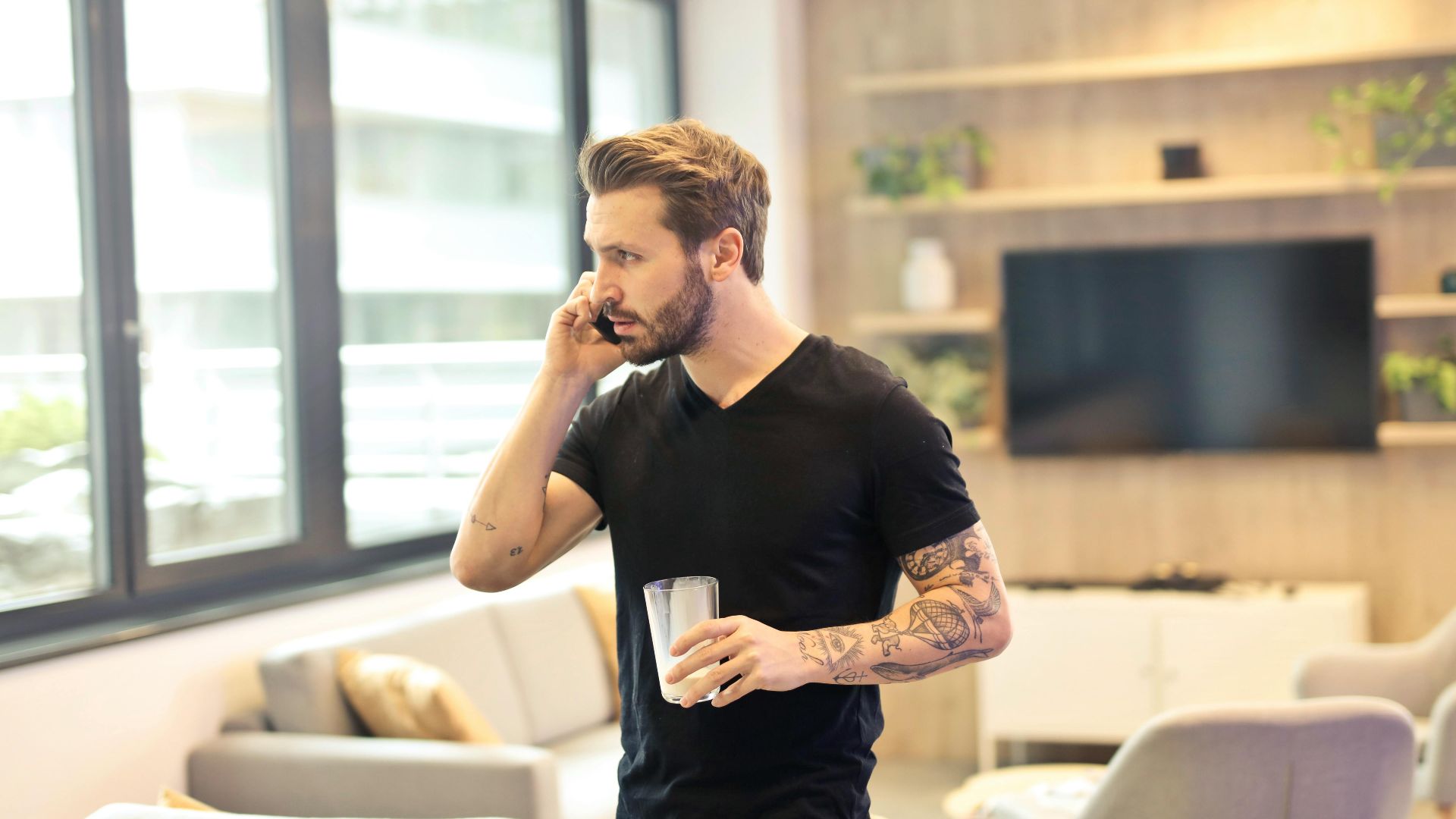 A tattooed man talking on a smartphone and holding a drink in a contemporary living room setting.