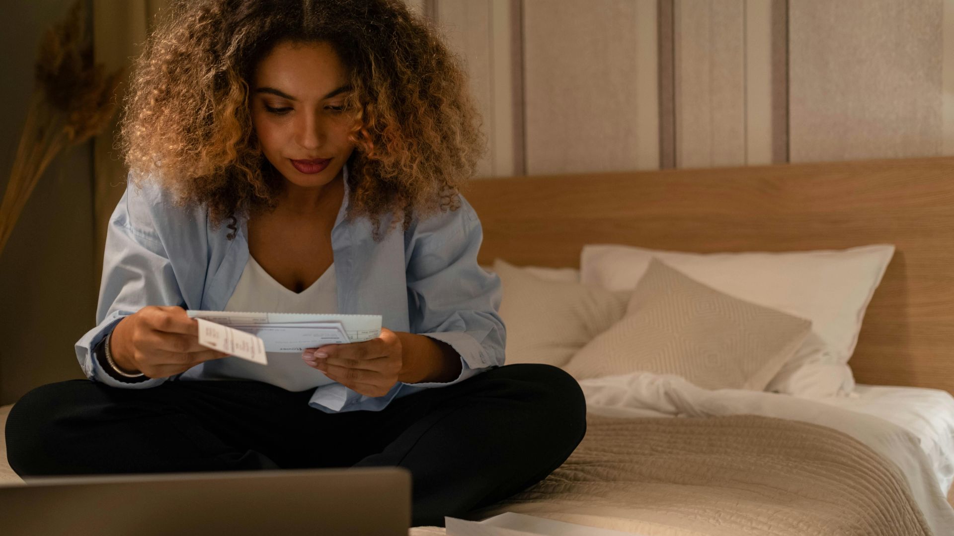 Woman sitting on bed reading documents with laptop, highlighting home office setting.