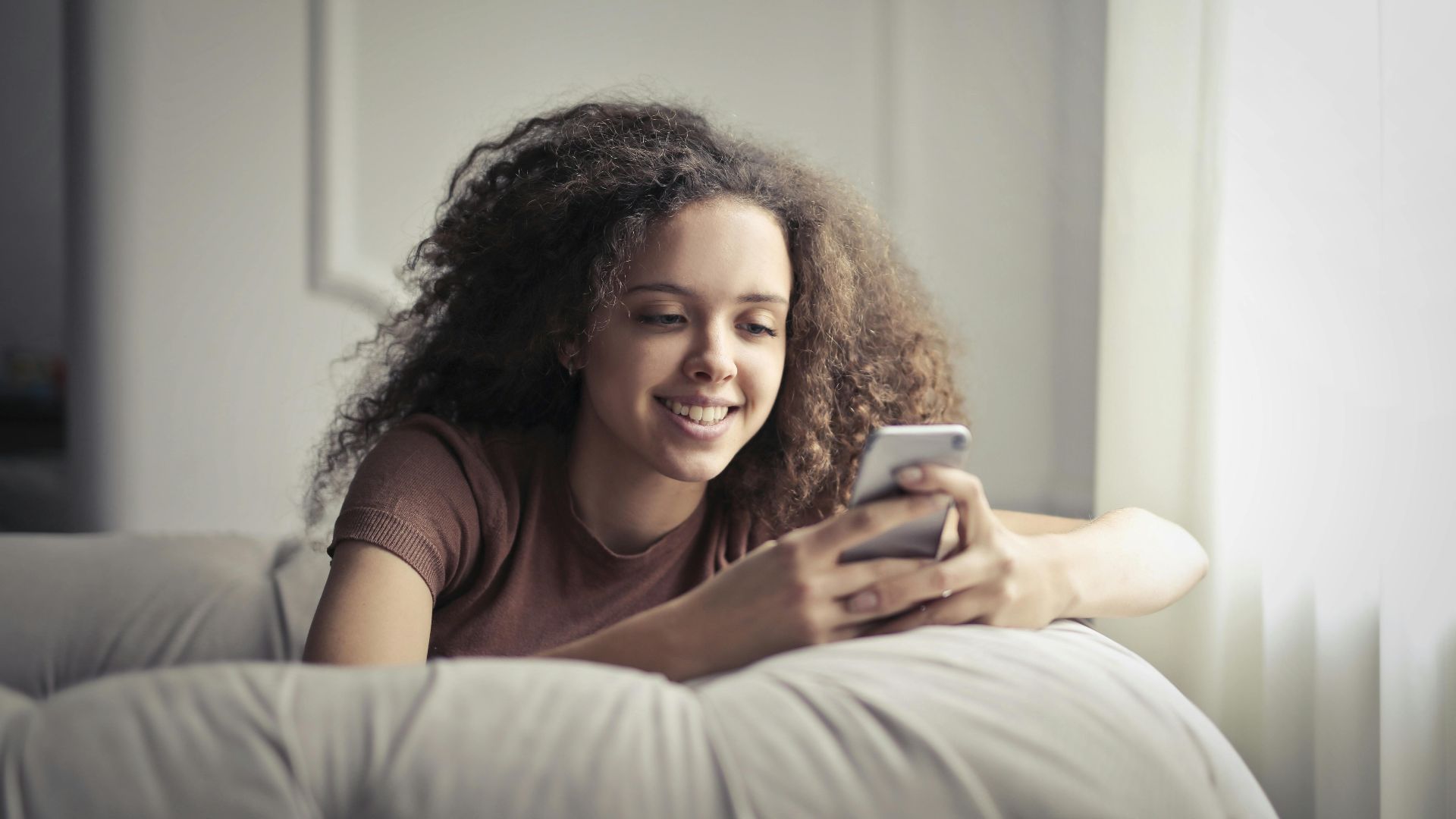 A smiling young woman with curly hair, comfortably texting on her smartphone indoors.