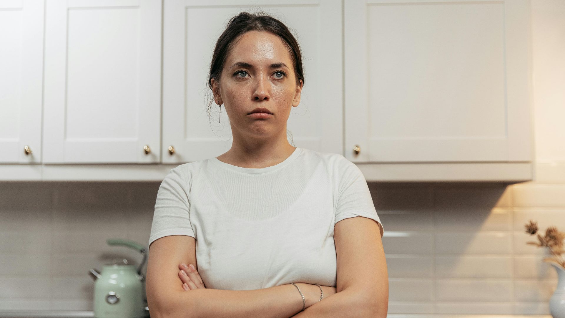 A woman in a white shirt crosses her arms, expressing discontent in a modern kitchen.