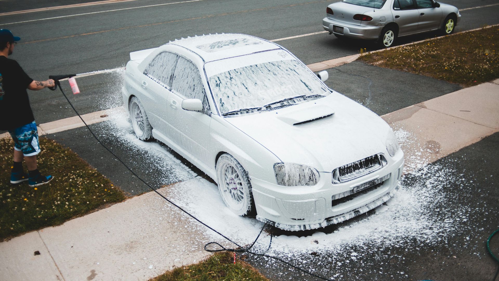 A man using a pressure washer to clean a white car with foam on a suburban driveway.