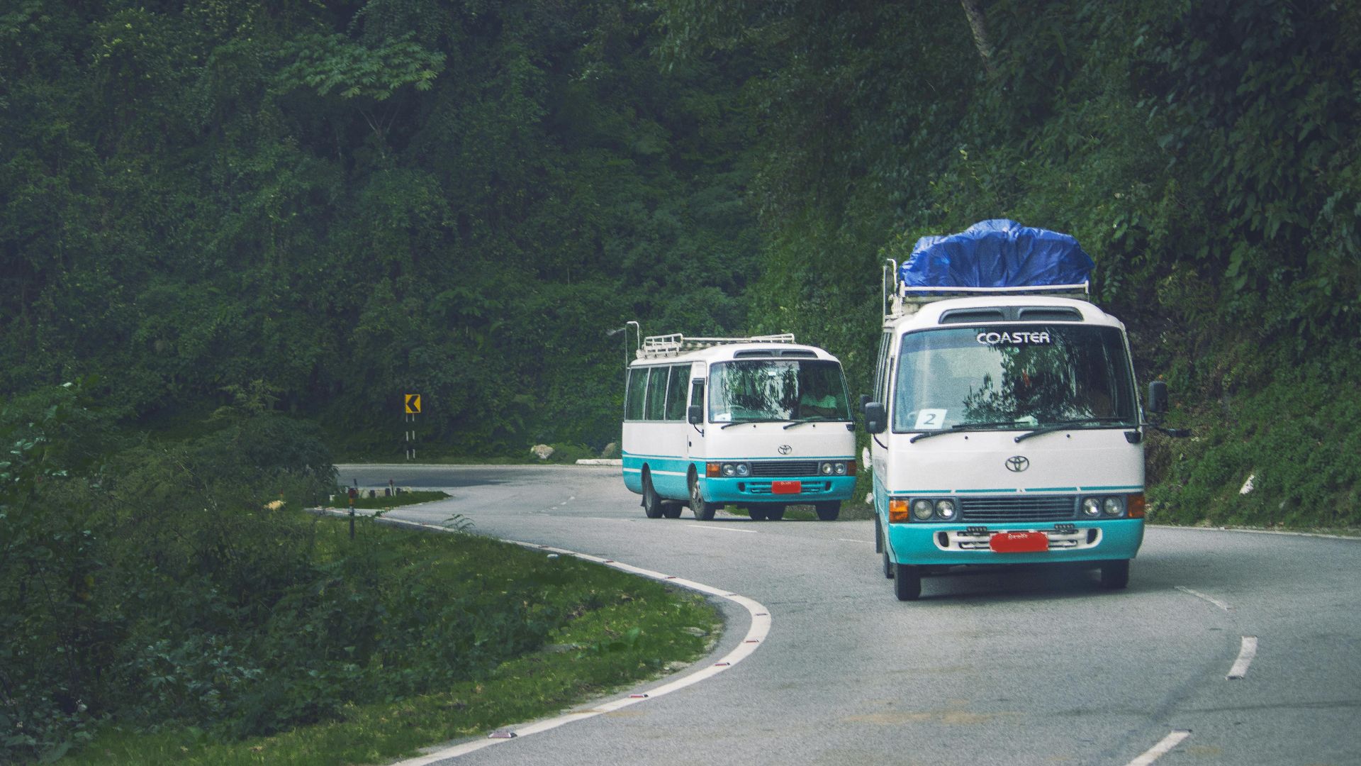 Two mini buses travel a scenic mountain road in Phuentsholing, Bhutan, surrounded by lush green forests.