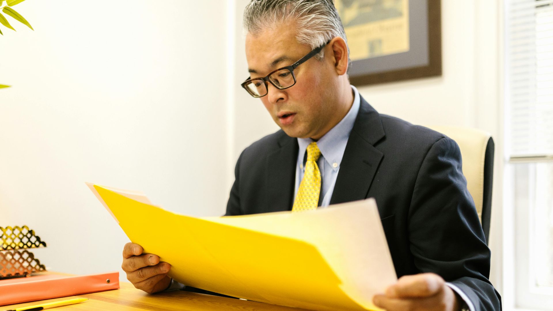 Asian businessman in corporate attire reading documents at office desk with a yellow folder.