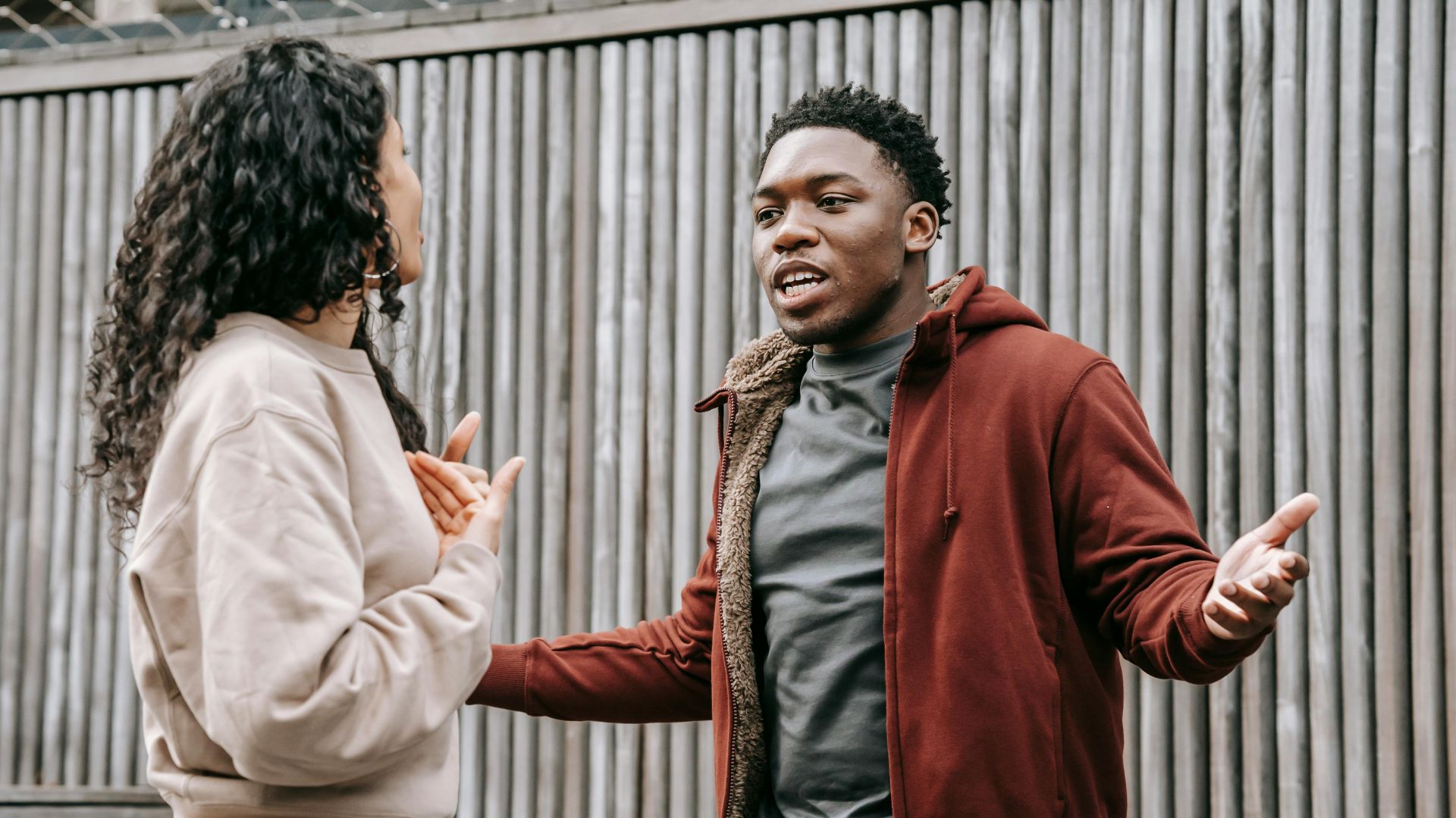 Angry African American boyfriend and ethnic girlfriend looking at each other while having emotional dispute on street near wooden fence