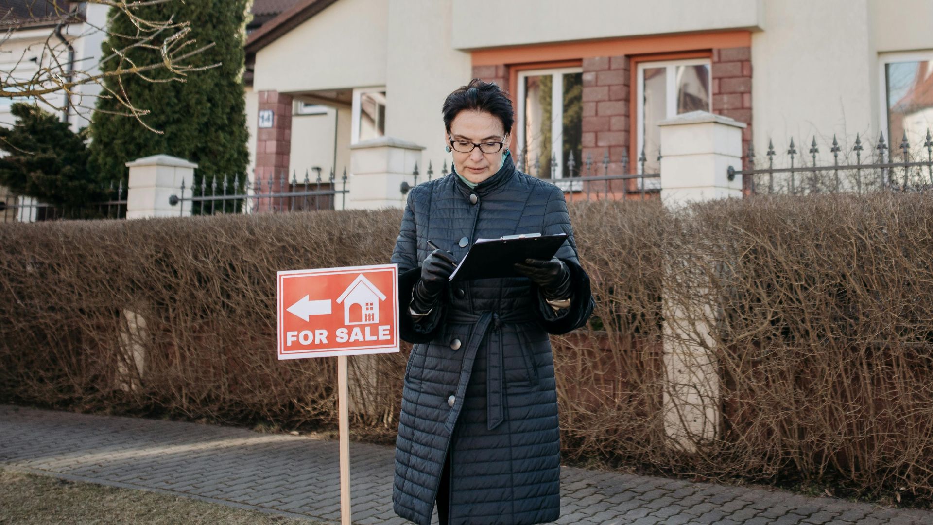 A real estate agent standing outside a house with a 'For Sale' sign, ready for viewing.