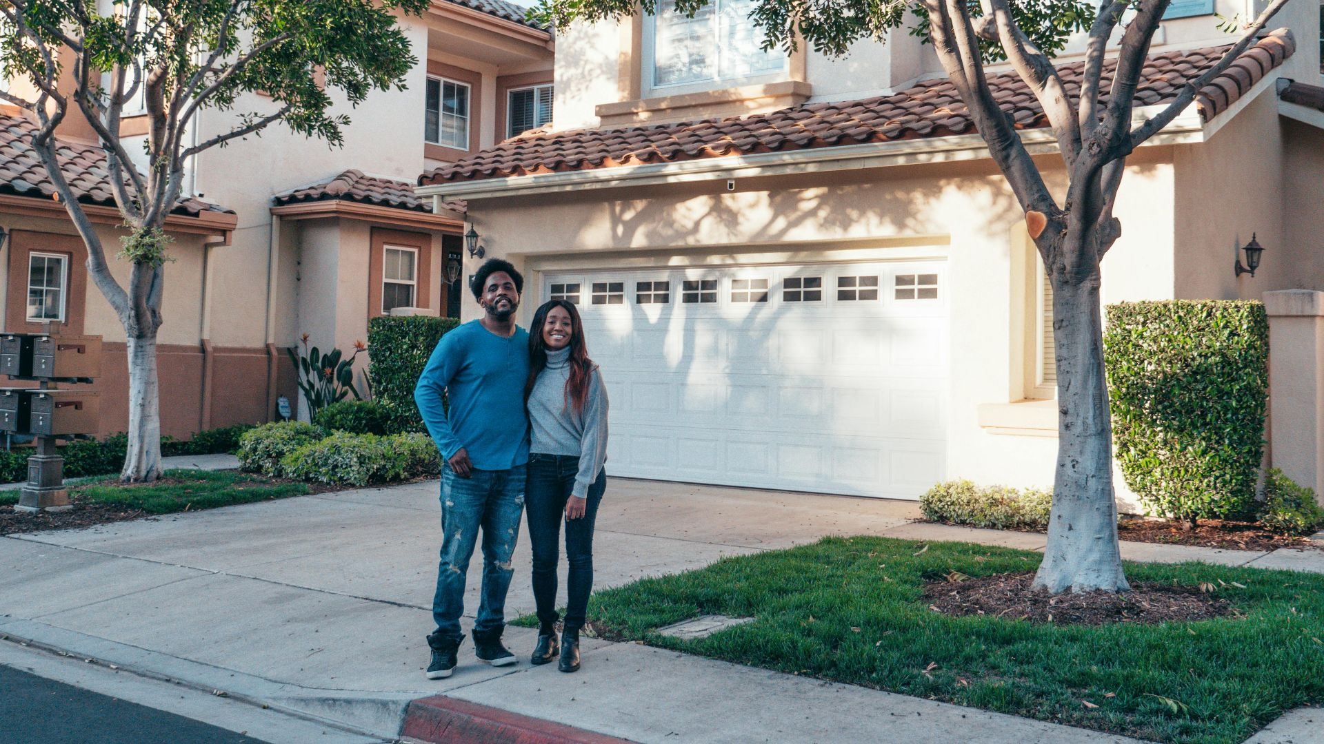 A smiling couple proudly posing in front of their newly purchased house, expressing joy and togetherness.