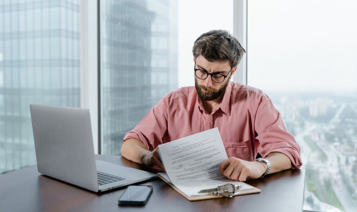 A Man in Pink Long Sleeves Wearing Eyeglasses while Looking at the Papers on the Table
