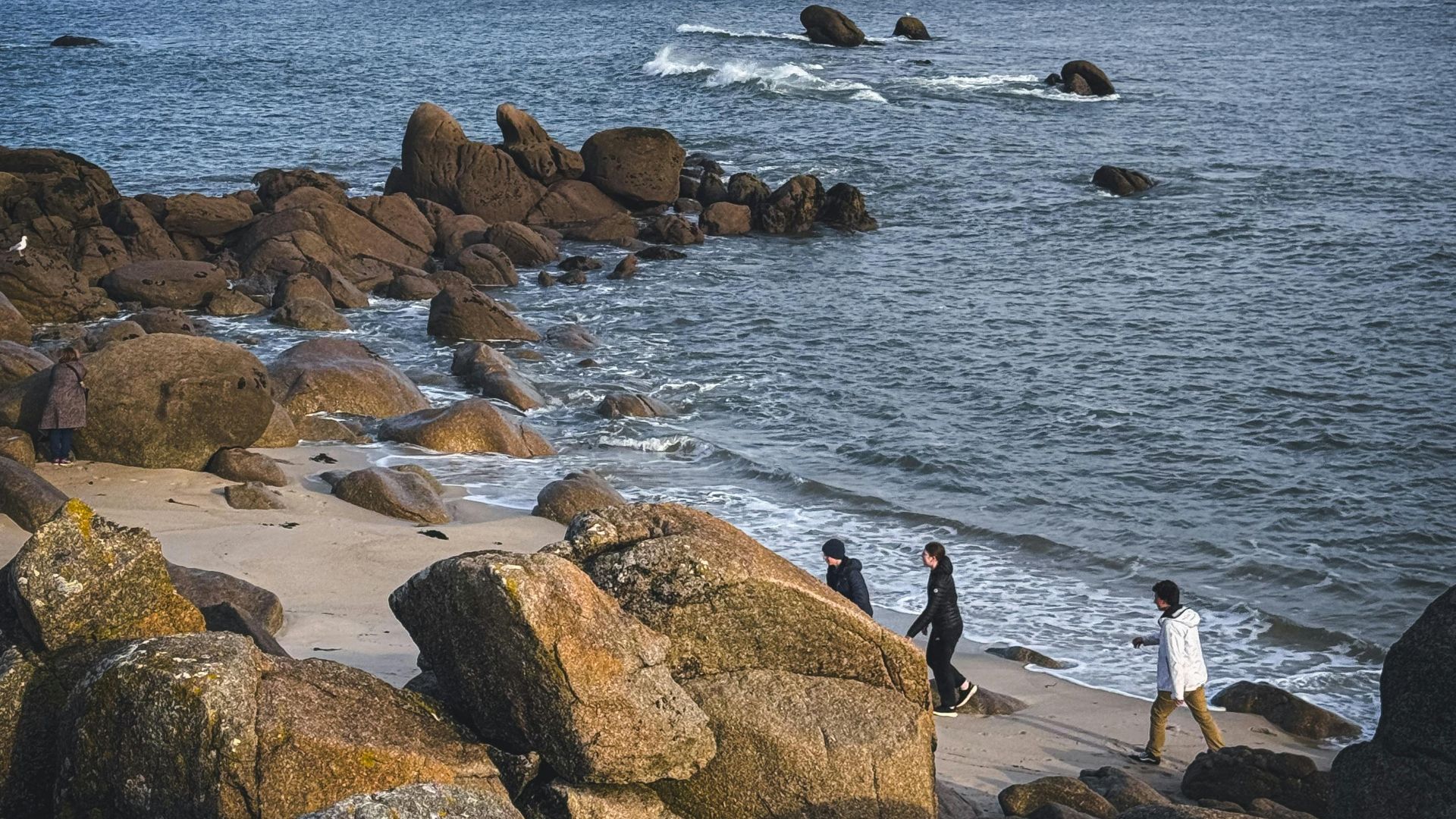 Three people walking by the rocky beach under a cloudy sky.