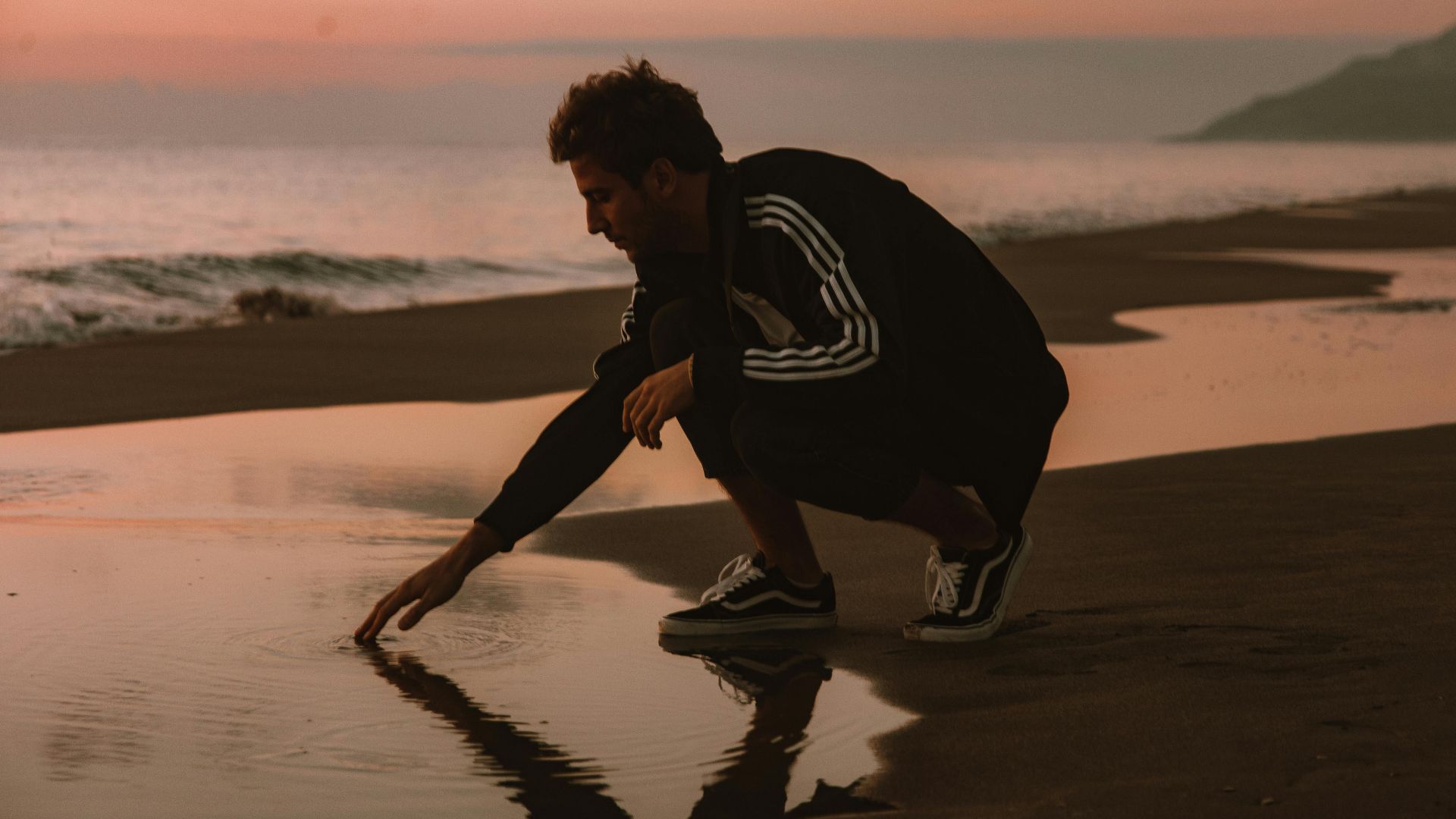 A man squatting at the beach during sunset, touching the sea reflection.