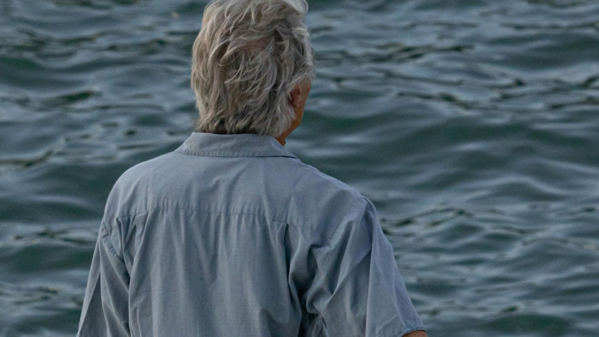 A man with gray hair stands relaxed by the sea, enjoying the summer breeze.