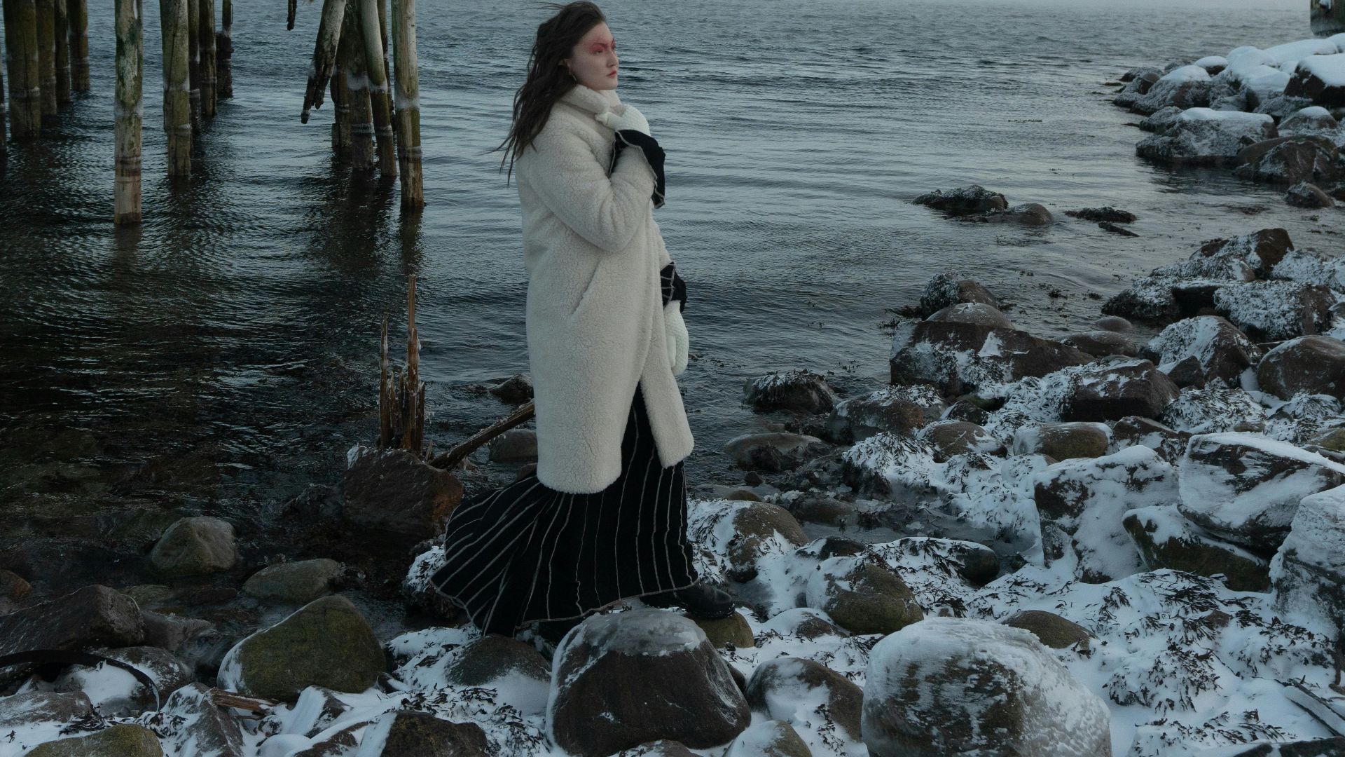 A woman stands in a fur coat near snowy rocks and a wooden pier by the ocean.