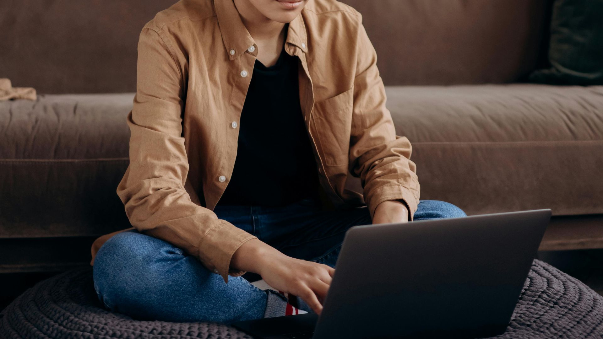 A woman sits on a floor cushion, working on a laptop in a modern living room.