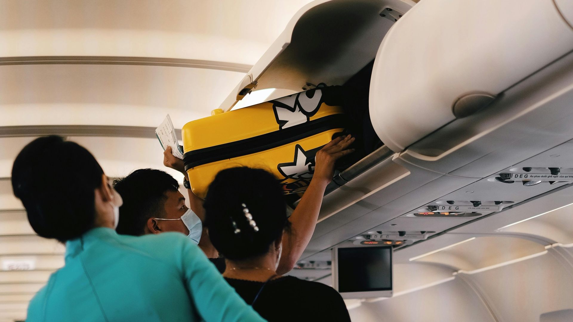 Passengers placing a yellow suitcase in the overhead bin of an airplane cabin. Interior with crew assistance.