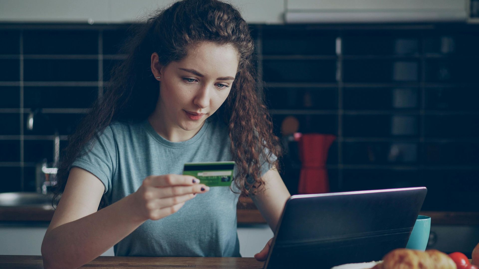 Young woman using a credit card to shop online with a laptop in a kitchen setting.