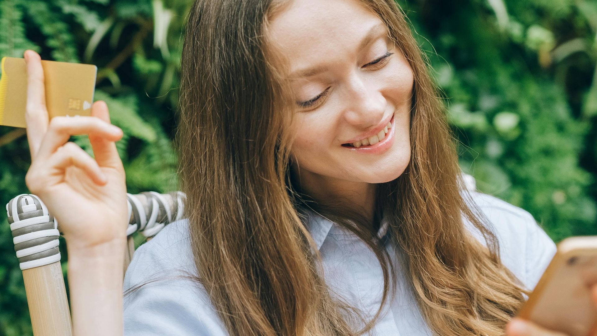Happy woman using smartphone and holding credit card in lush garden setting, enjoying online shopping.