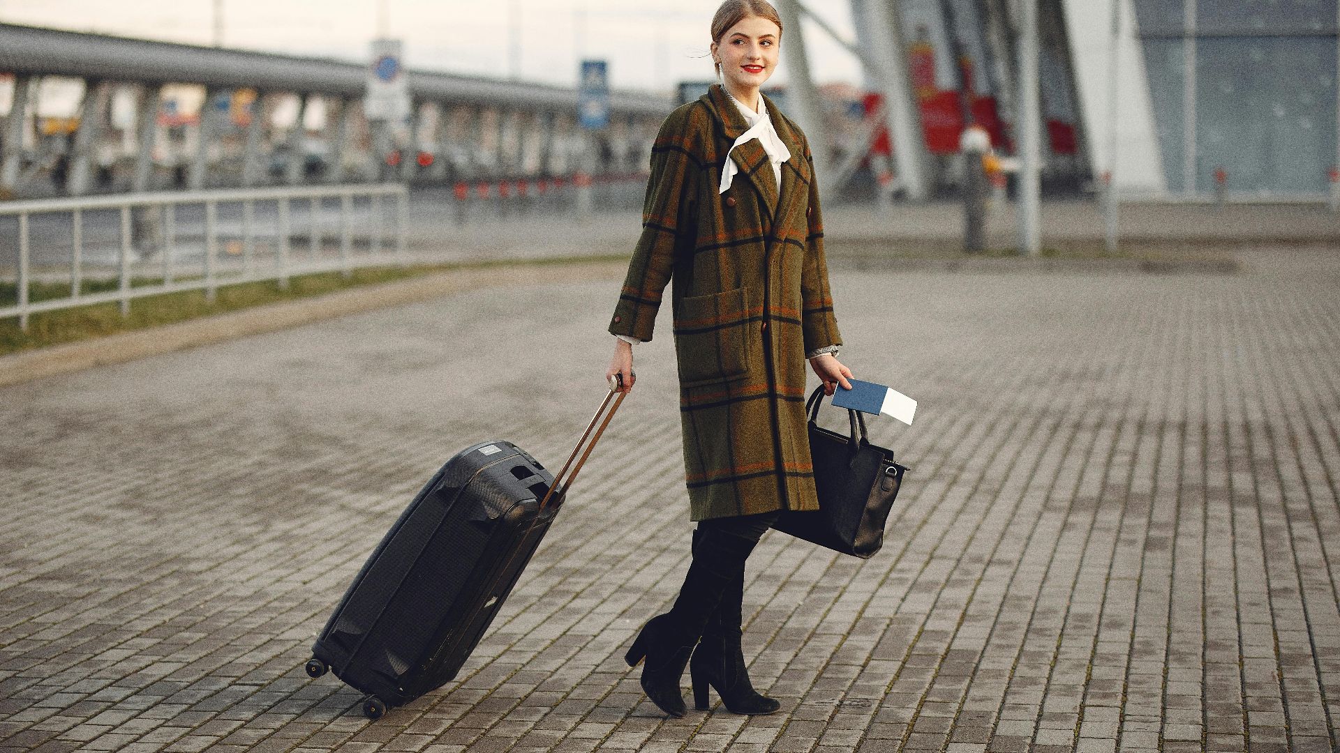 Full body young female passenger in trendy plaid coat with suitcase strolling on pavement near modern building of ariport terminal taking bag and documents
