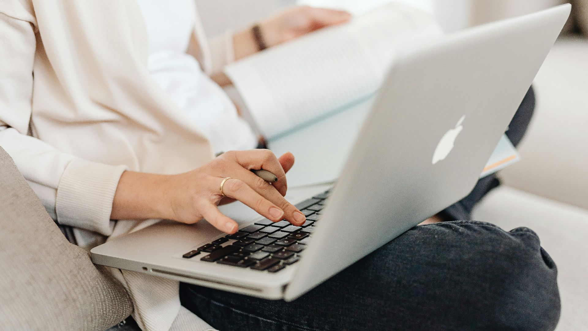 Close-up of a person multitasking, typing on a laptop while holding a book on their lap.