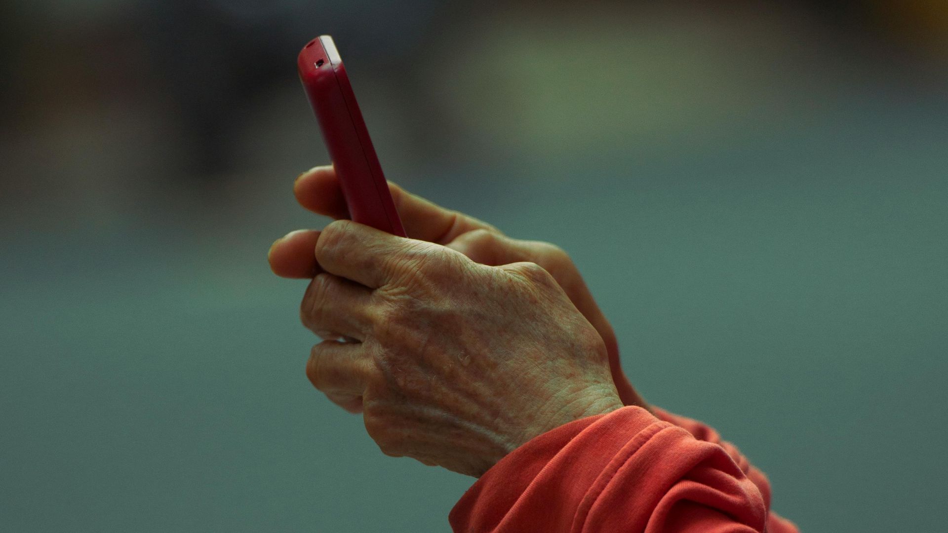 Close-up of hands using a smartphone in a red case outdoors. Focus on device, blurred background.