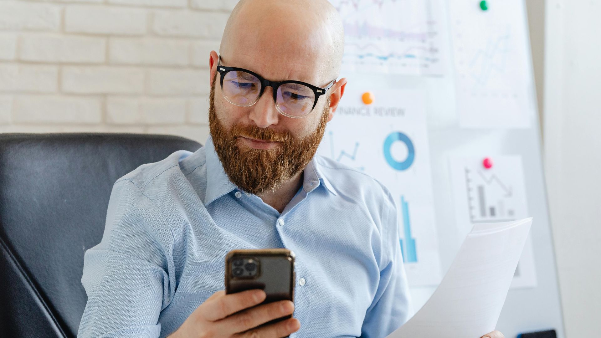 Bald businessman with eyeglasses checking phone data during an office presentation.