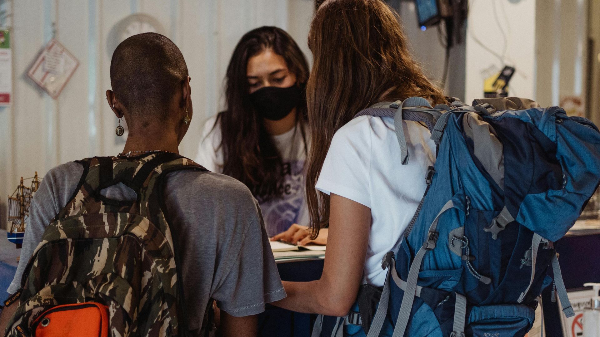 Two backpackers checking in at a hostel reception, wearing face masks.