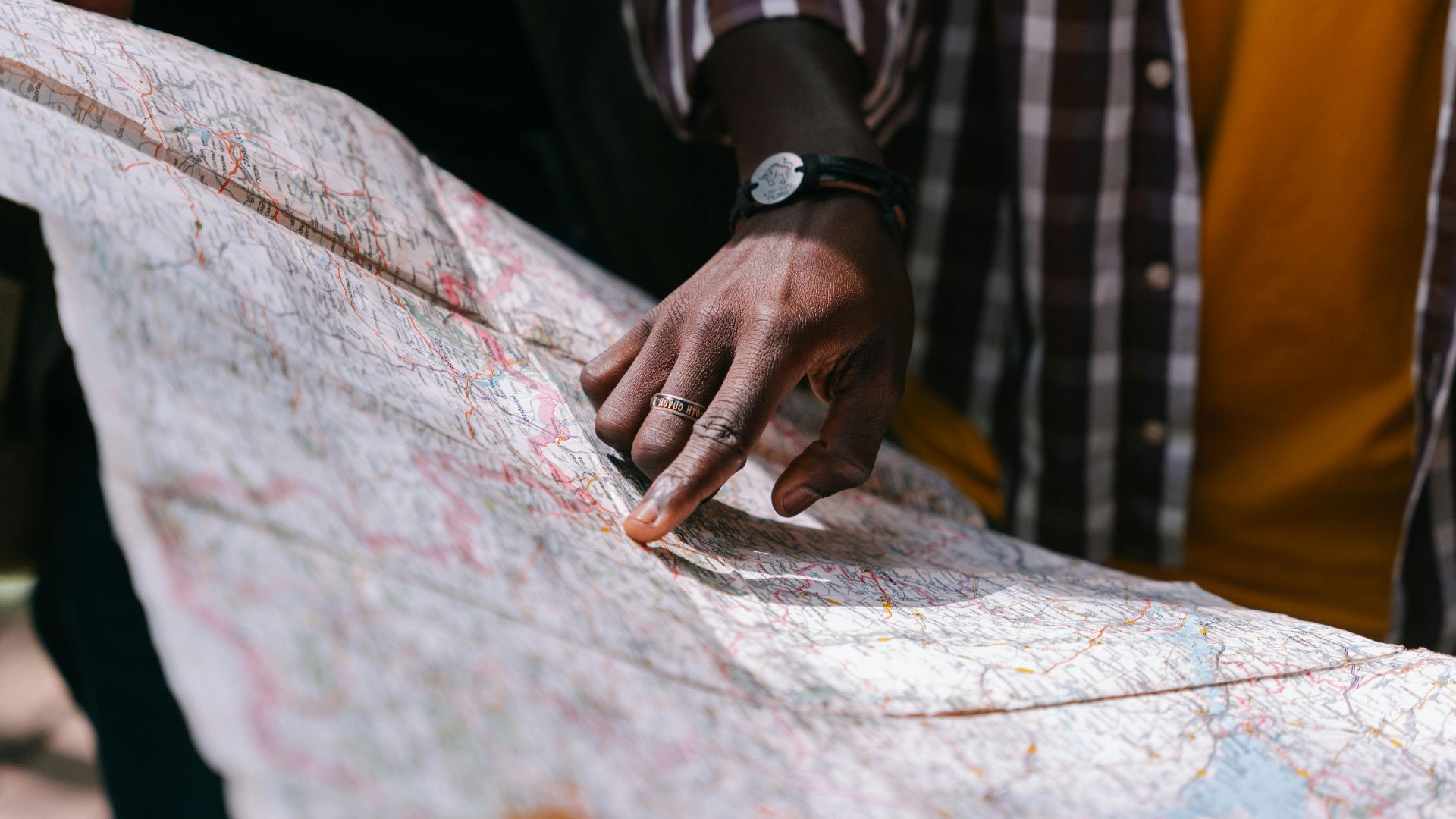 Close-up of a hand pointing at a detailed map, outdoors, wearing a striped shirt.