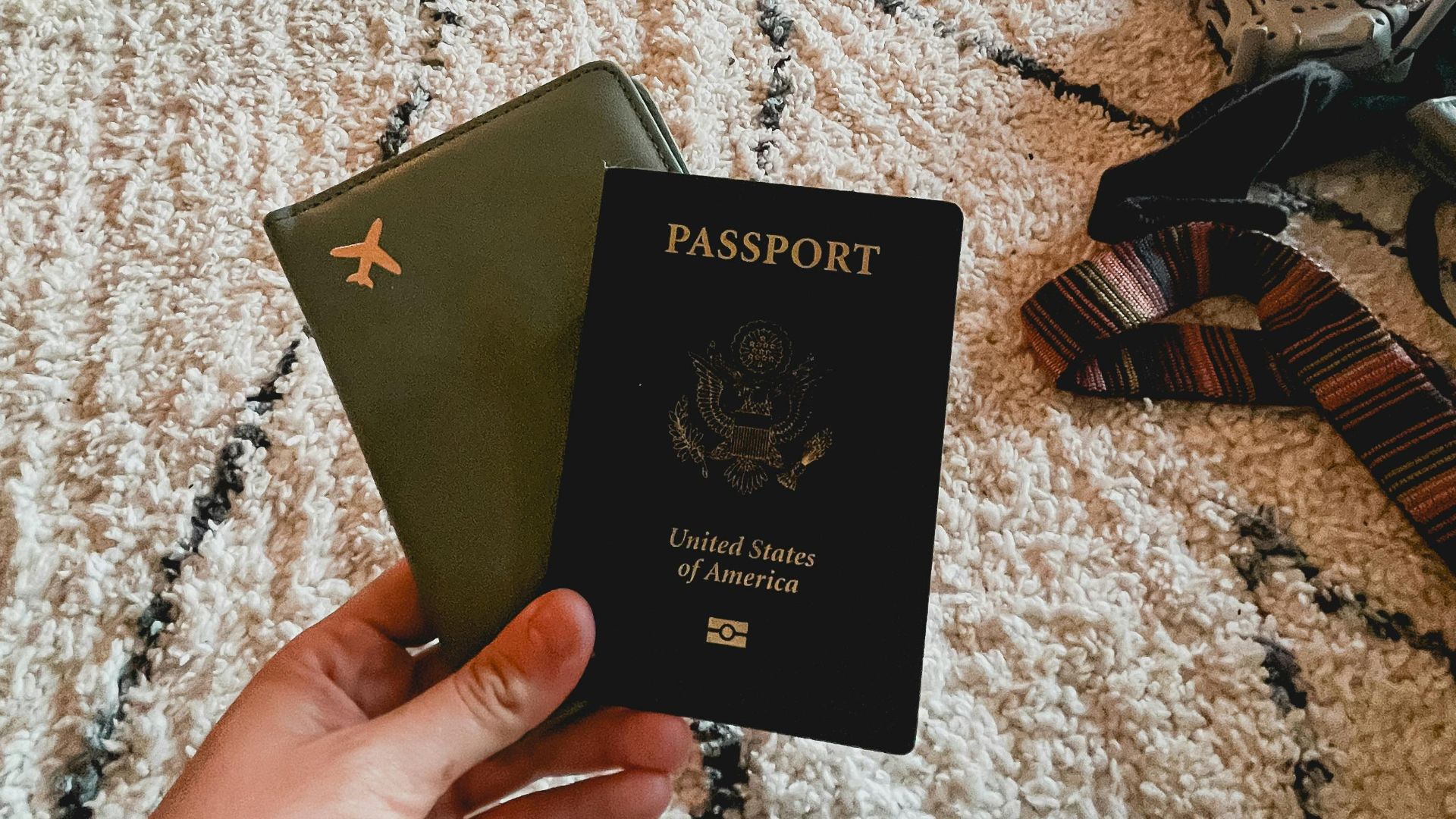 Hand holding two passports on a patterned carpet indoors, travel essentials.