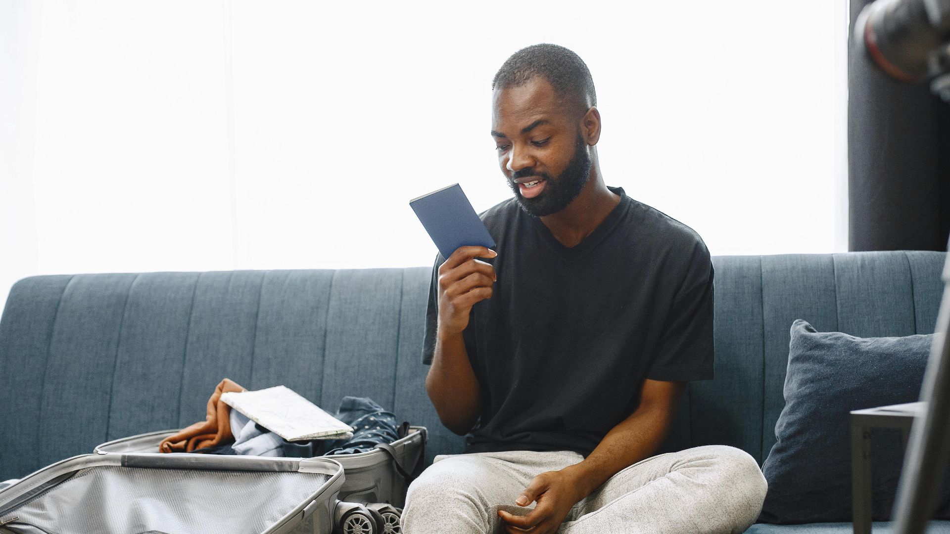 Adult man sits on sofa with open luggage, holding passport, preparing for travel, vlogging.