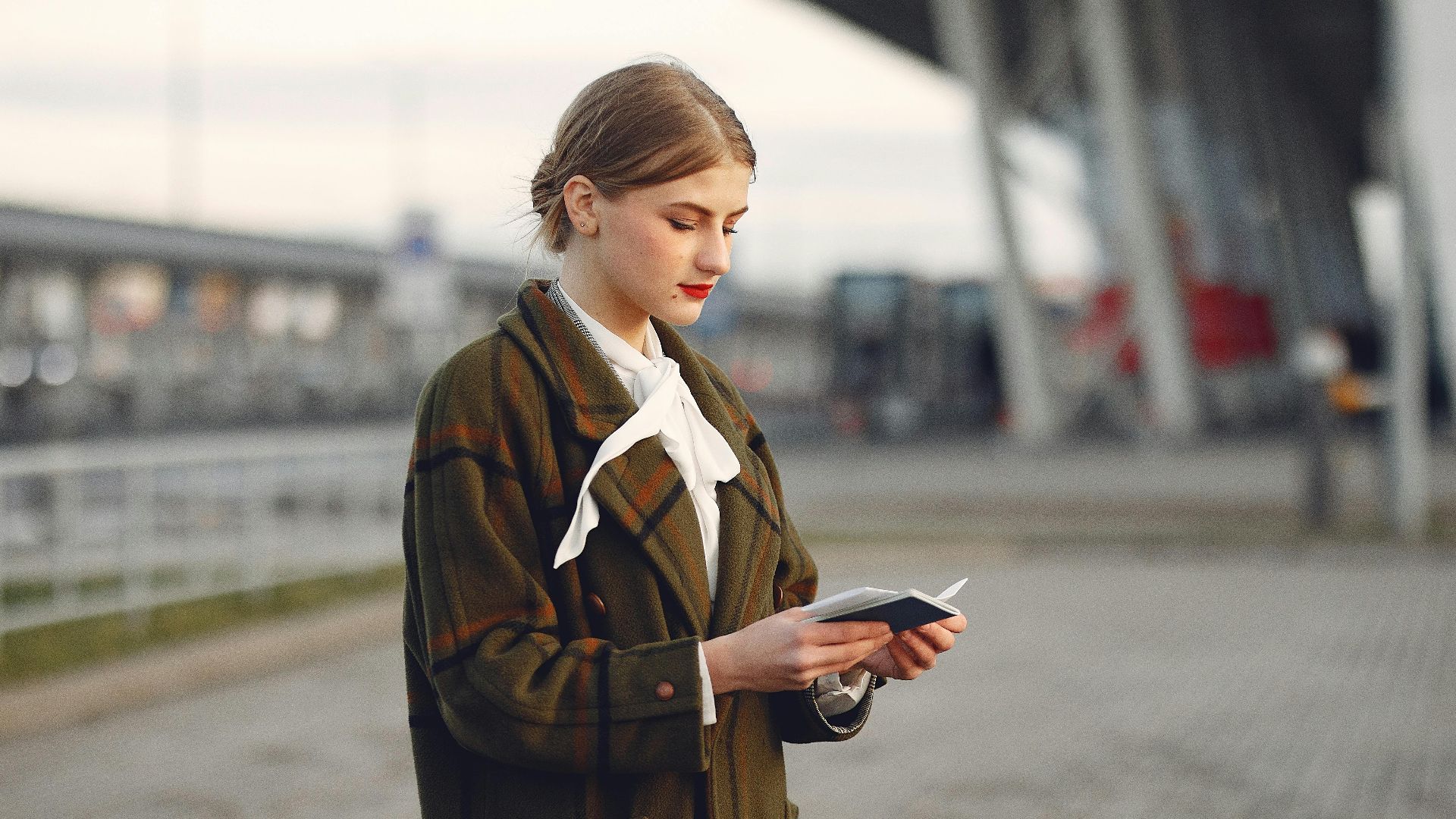 Attentive female passenger wearing trendy plaid coat and white blouse checking passport and ticket standing on pavement near modern building of airport outside