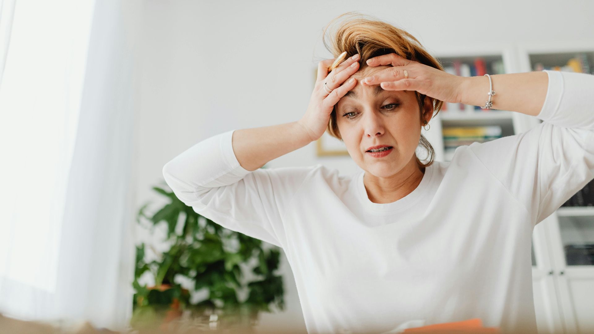 A worried woman indoors with hands on forehead, expressing stress.