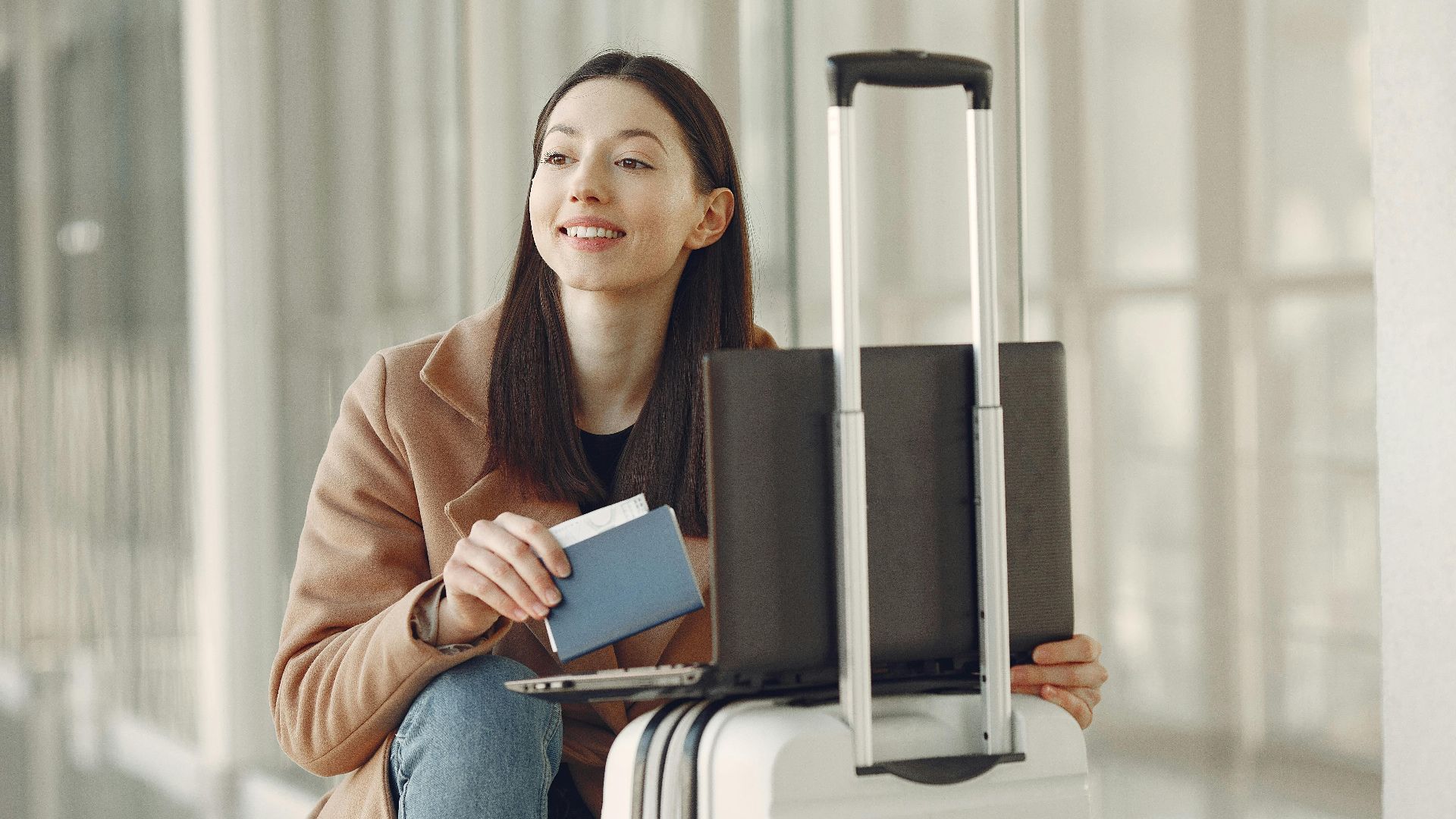 Smiling adult ethnic female traveler in trendy coat holding passport and using laptop on luggage in airport corridor for checking ticket for correctness