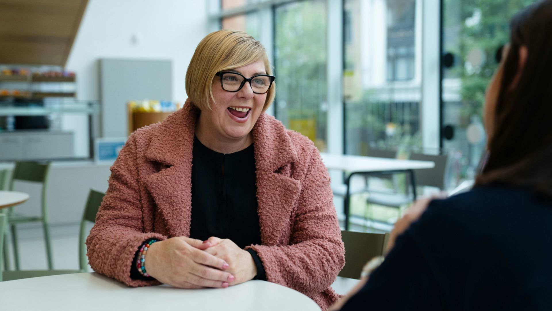 Two people talking at a table by large windows.