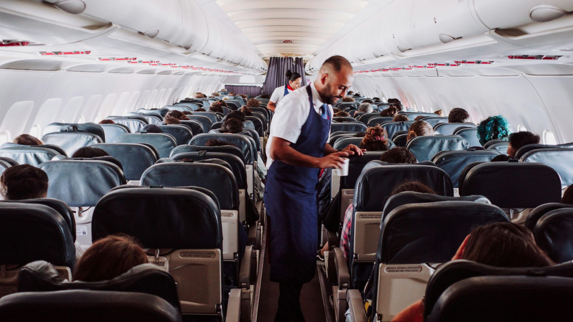 Flight attendant assists passengers in a crowded airplane aisle with rows of seats.