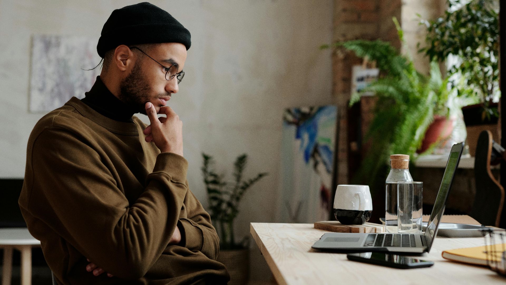 A man in casual attire working thoughtfully on a laptop in a cozy home setting with plants.