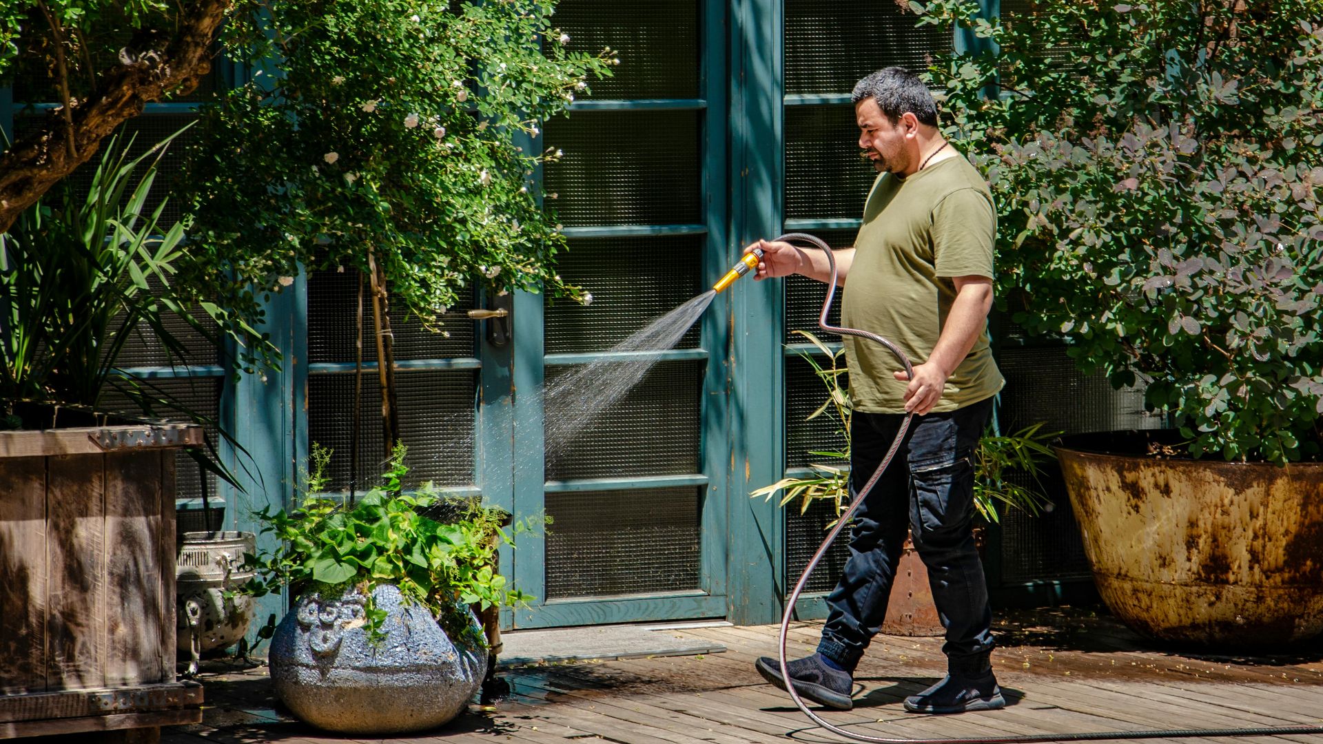 A man attentively waters plants on a sunny garden patio, emphasizing outdoor care and greenery.
