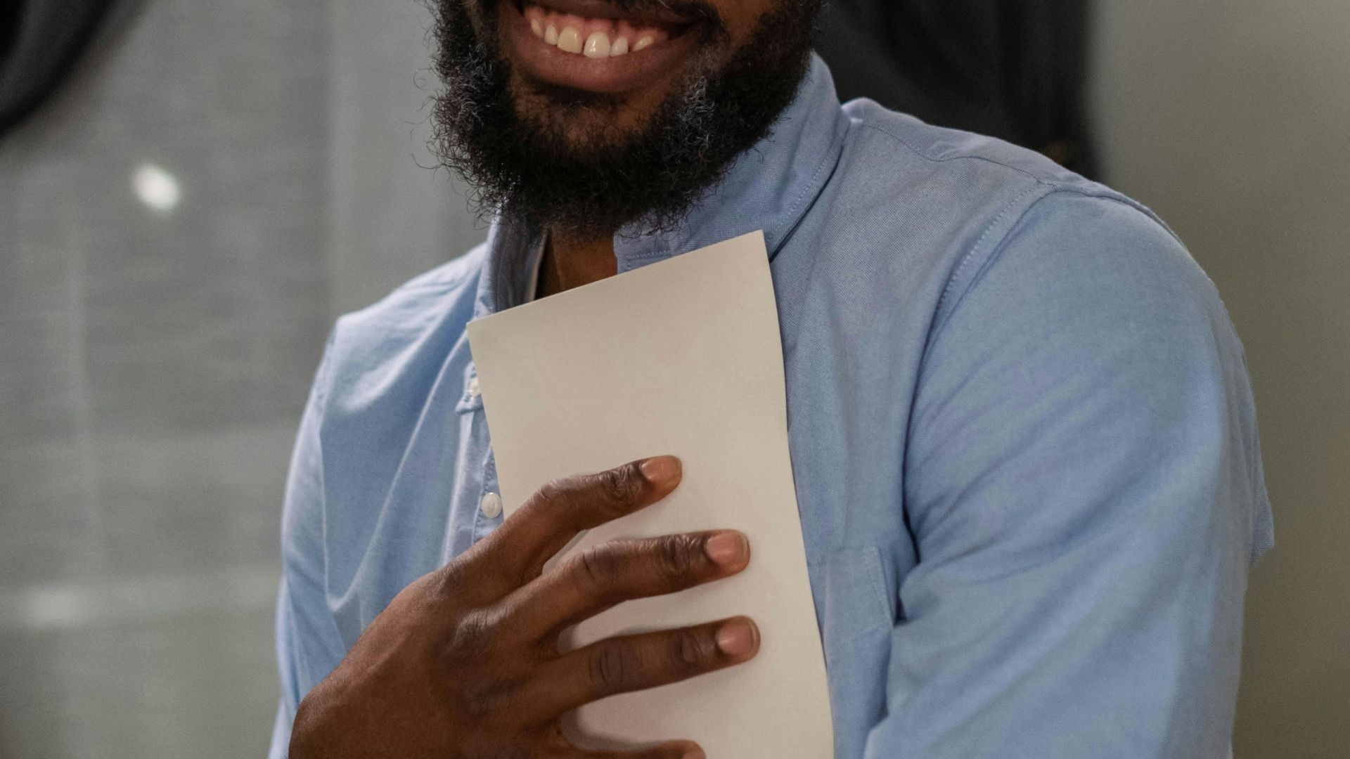 A happy man with glasses holding a paper, smiling indoors with natural light.