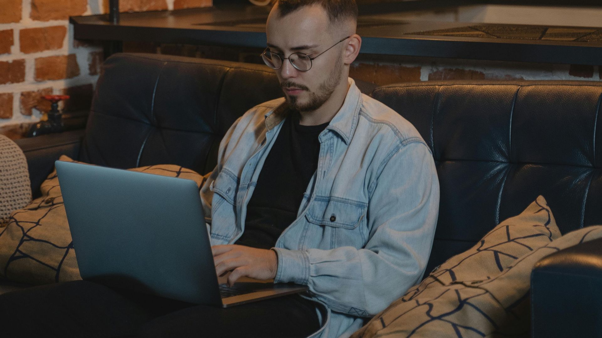 Bearded man in denim jacket using laptop on couch in industrial-style living room, working remotely.