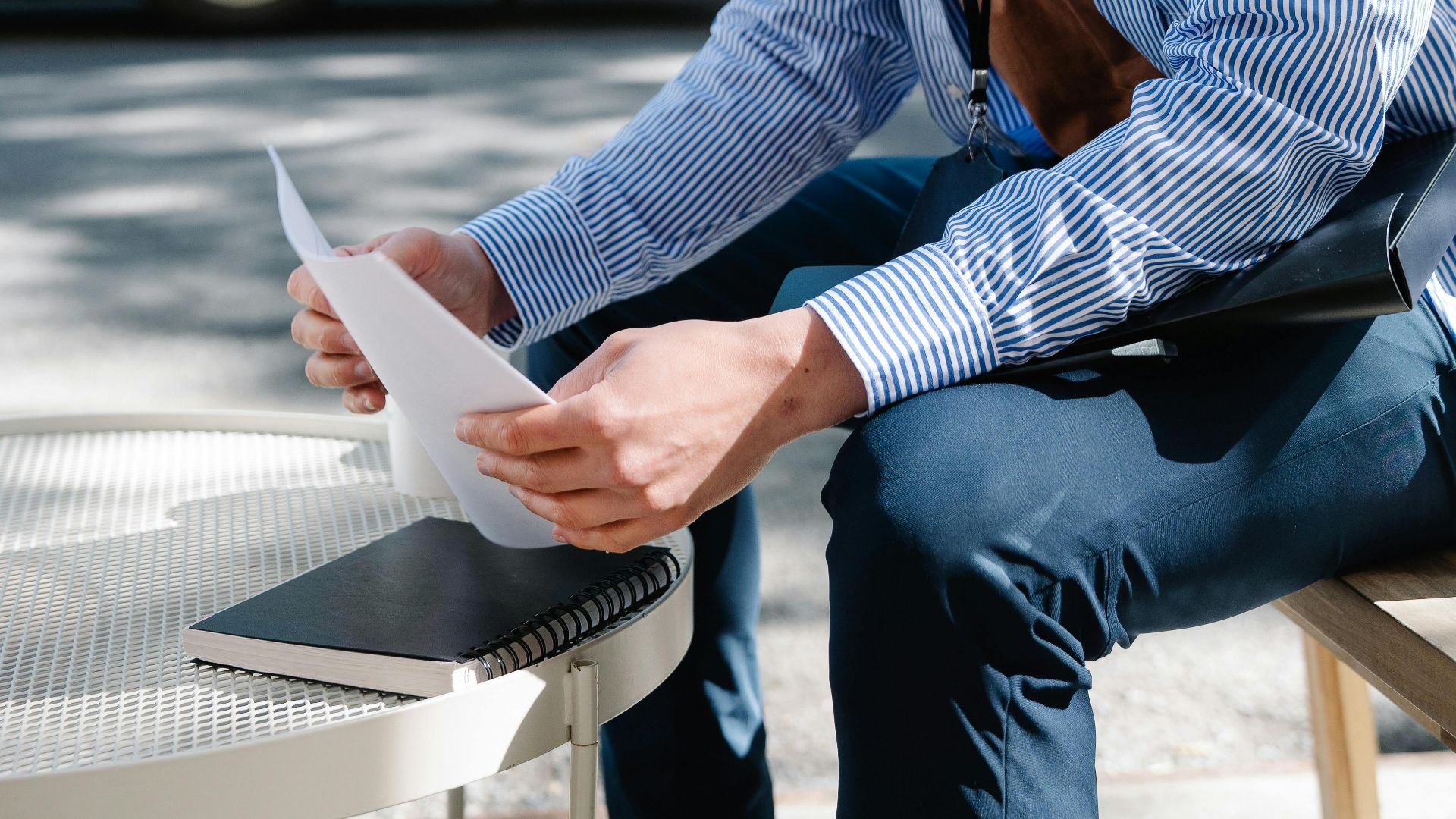 Asian man in striped shirt sitting outdoors reviewing documents at a table.