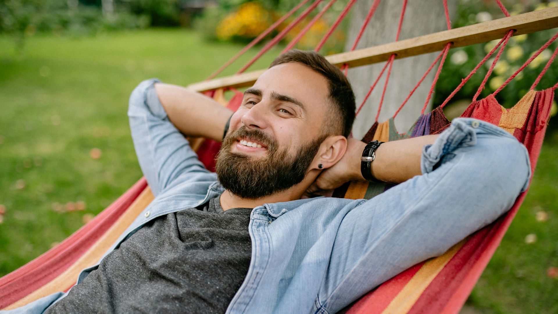 A bearded man enjoys a sunny day relaxing in a hammock in a garden.