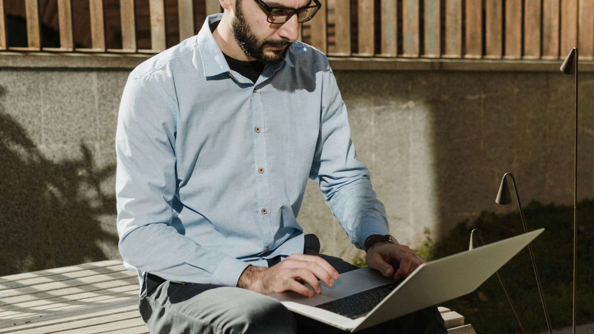 Caucasian man sitting outdoors working on laptop in sunlight with casual attire and eyeglasses.