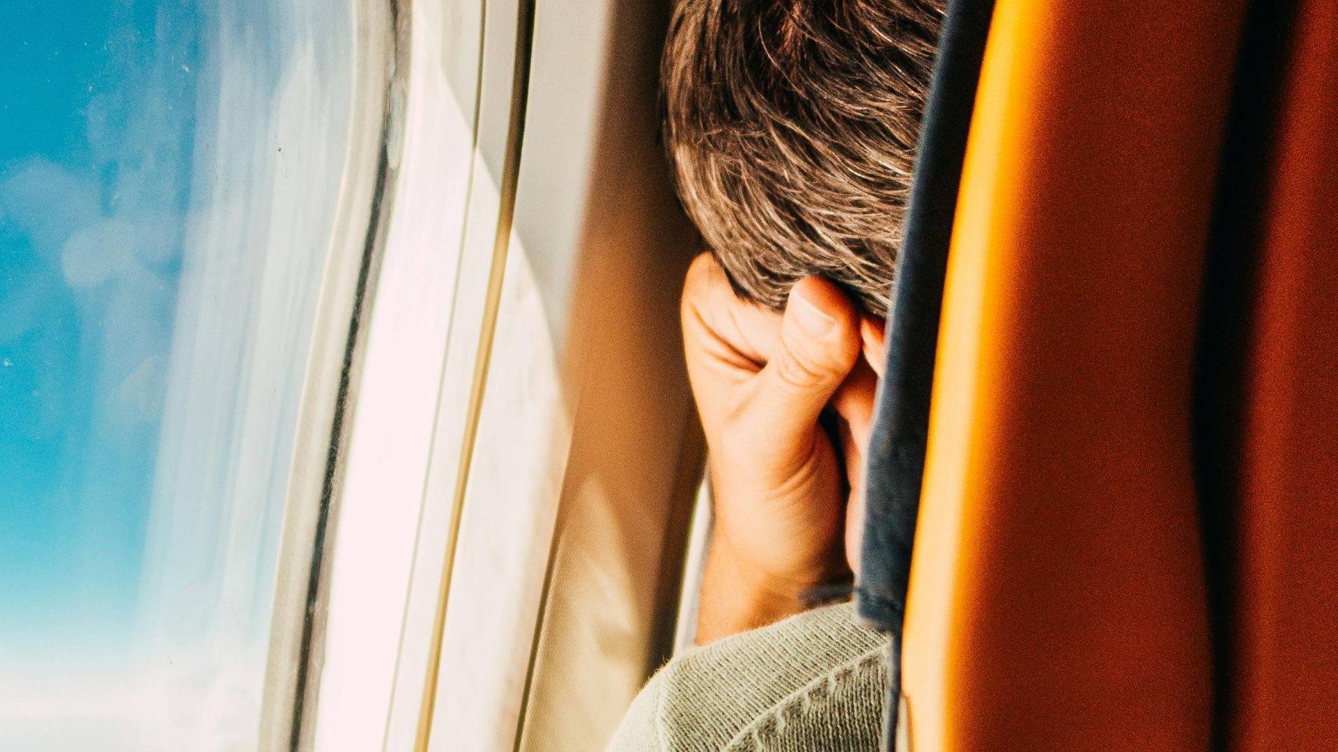 A man resting by the airplane window, peering into the sky during a daytime flight.