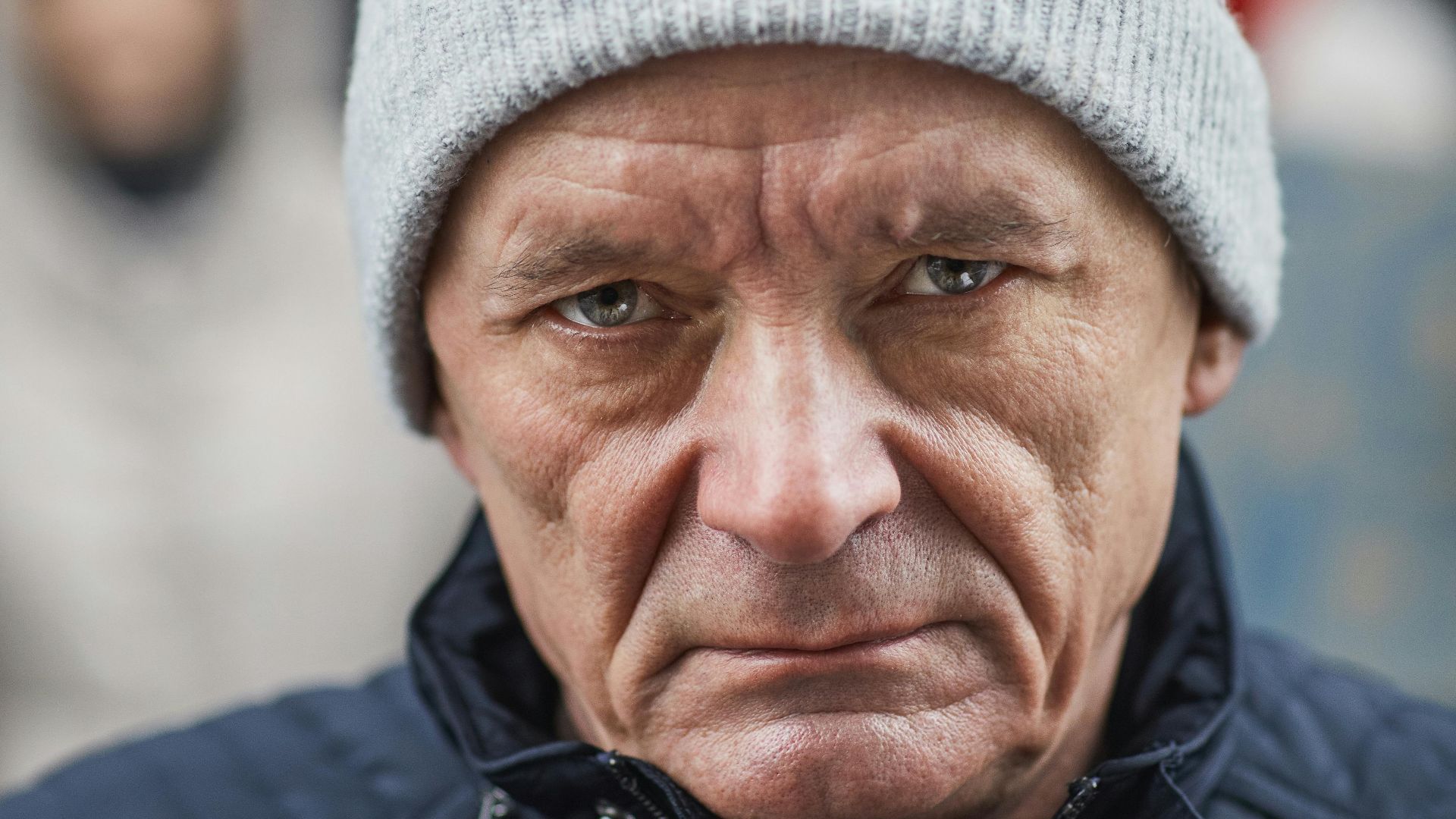 Close-up of a senior man in a beanie, looking serious and holding a handheld device, possibly a microphone.