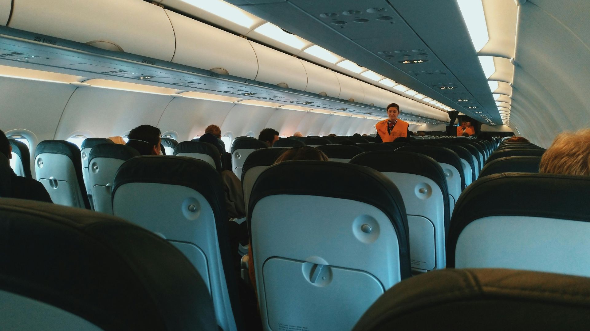 Inside of modern airplane cabin with passengers sitting on comfortable seats and cabin crew standing at passageway
