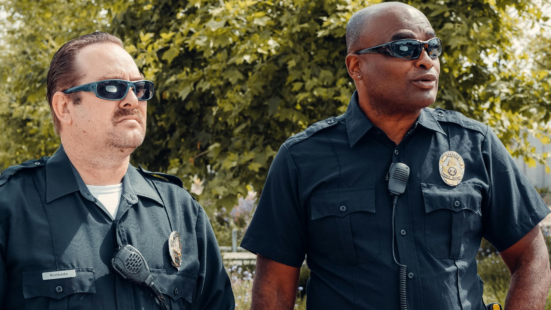 Two police officers in uniform, wearing sunglasses, standing outdoors with a serious demeanor.