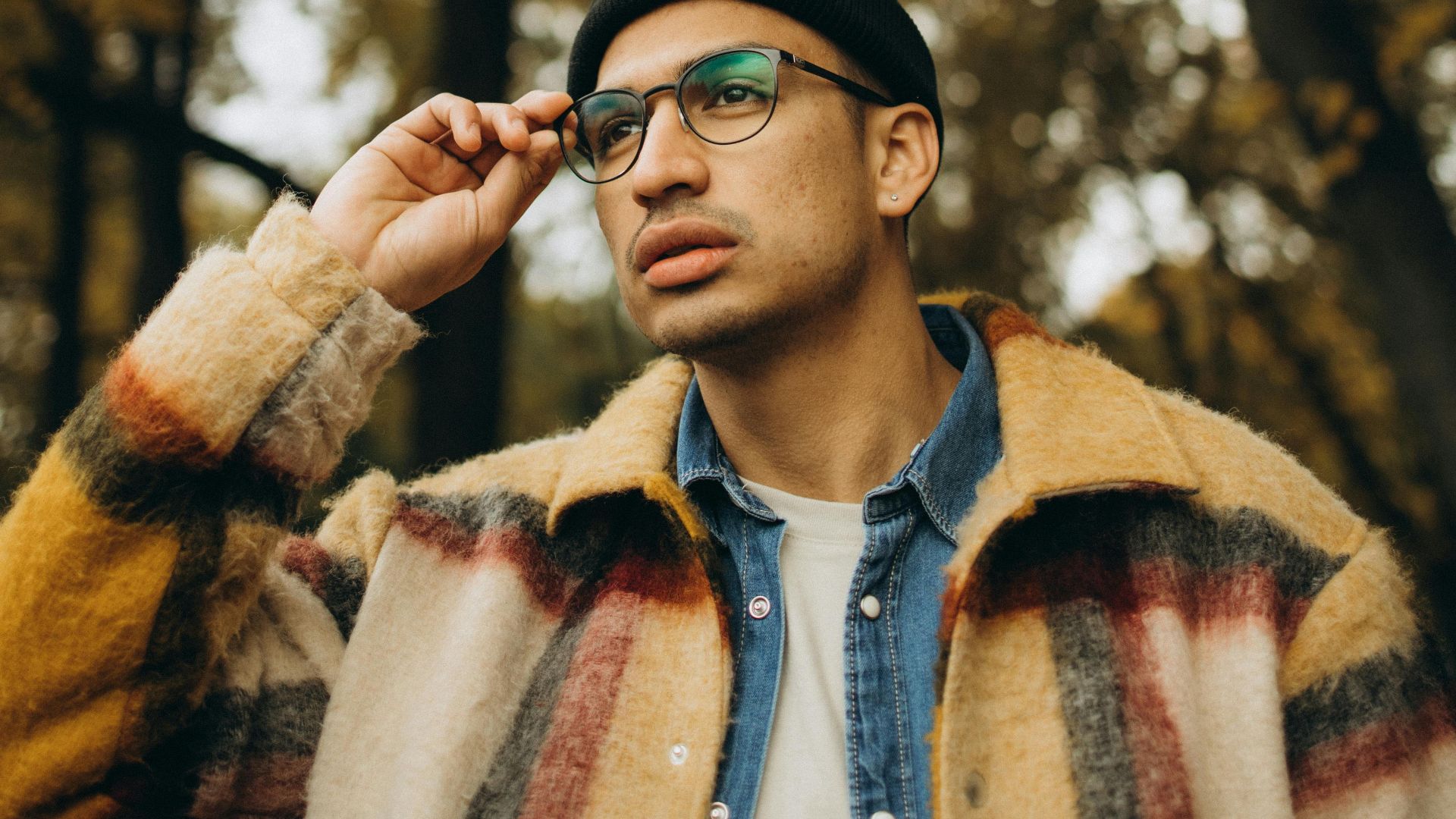 A fashionable man with eyewear and a checkered coat thoughtfully posing in a serene fall setting.