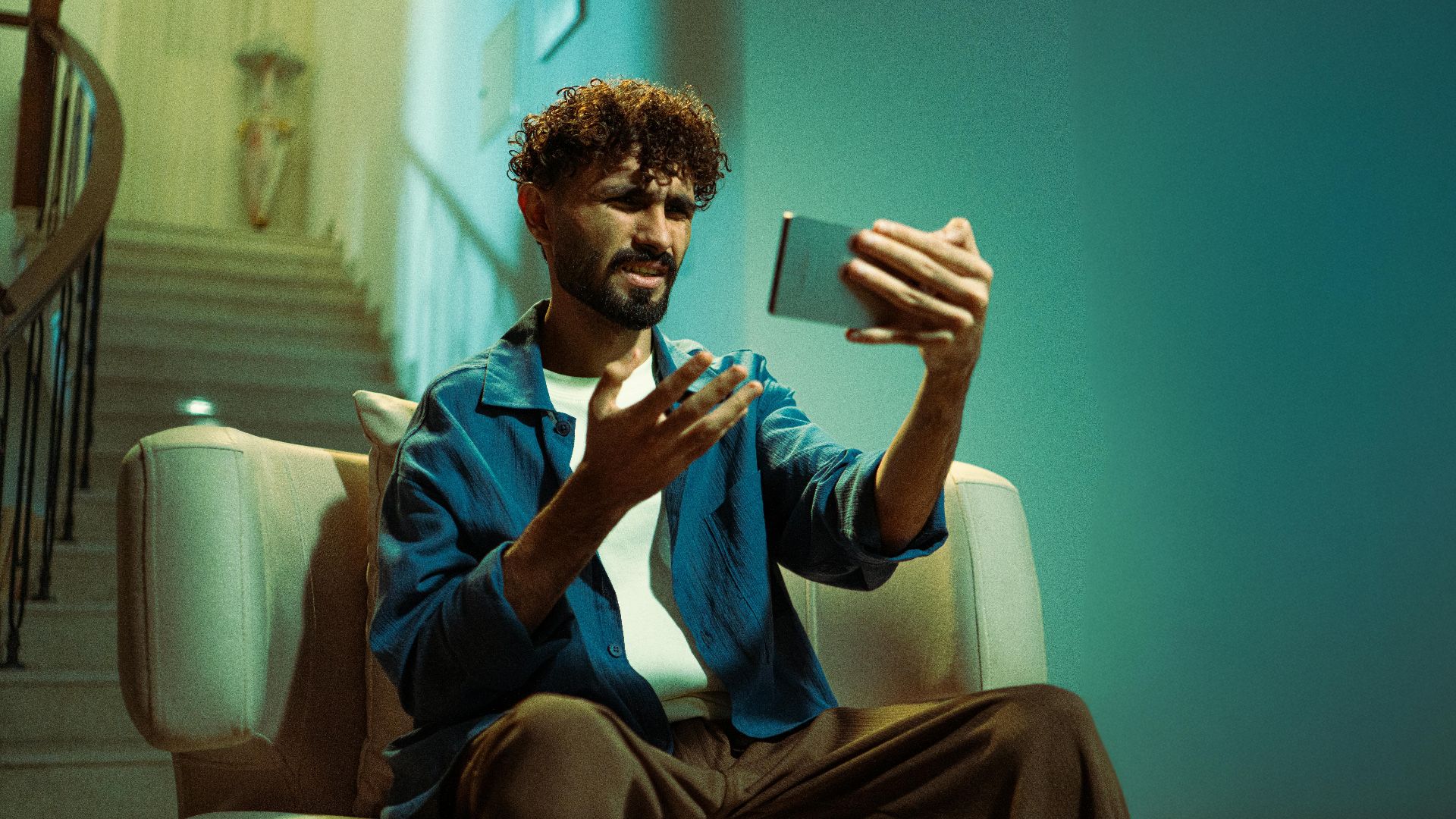 A man sitting on a couch indoors, looking confused while using a smartphone.