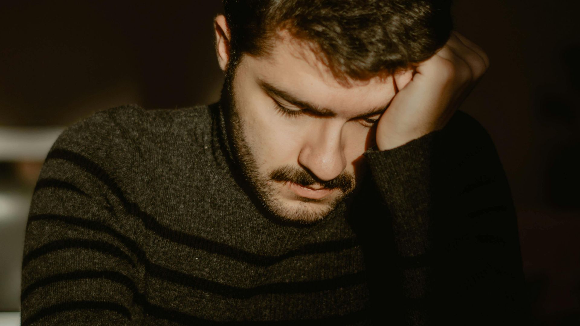 Exhausted ethnic male with mustache sitting at wooden table while looking down in evening in house